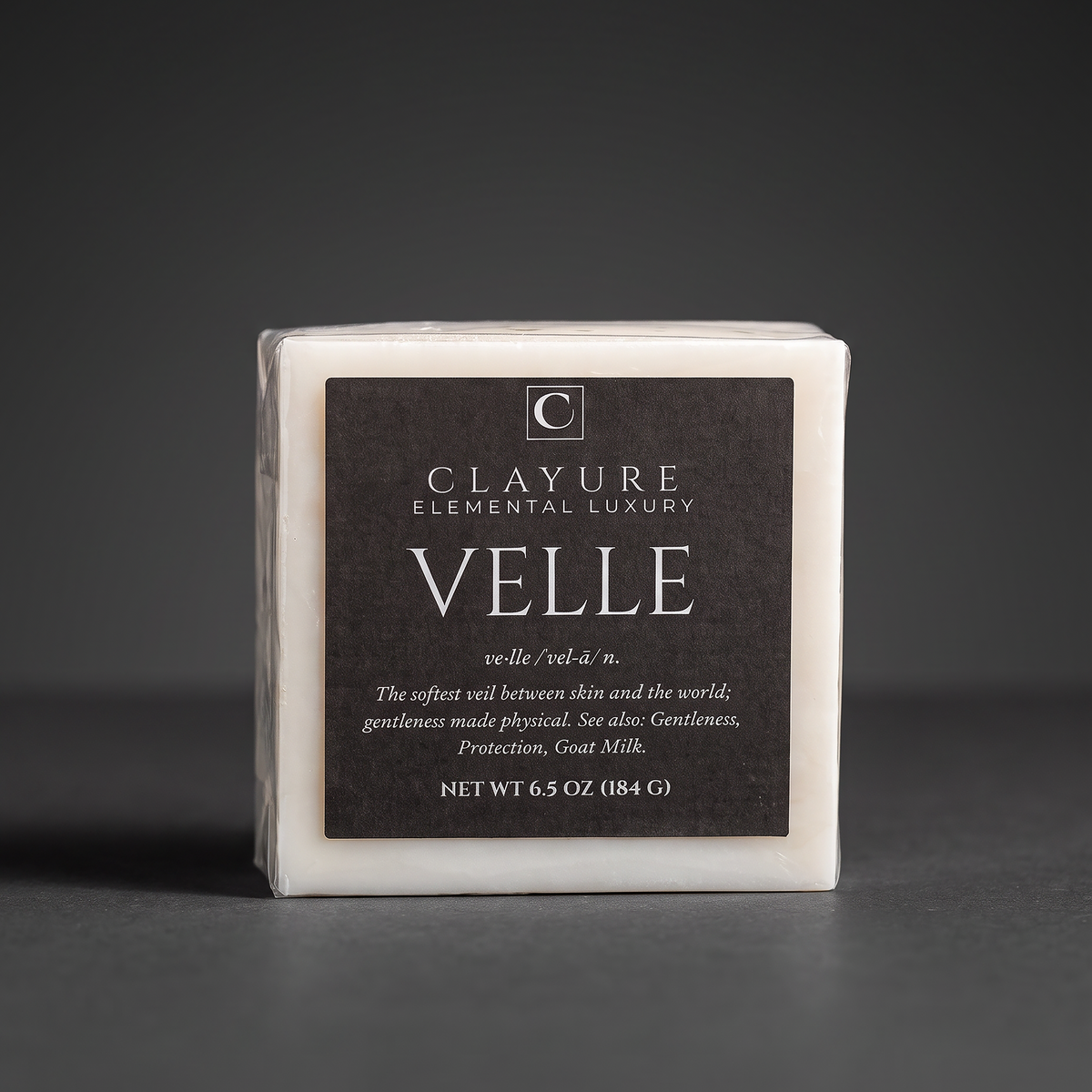 A square white bar of Clayure Velle goat milk soap wrapped in clear plastic with a large dark brown label, photographed on a dark gray background.