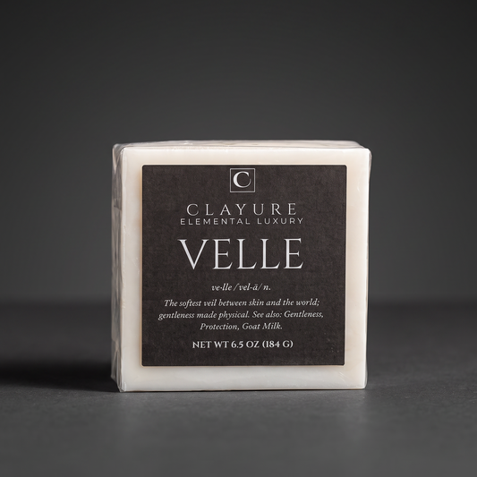A square white bar of Clayure Velle goat milk soap wrapped in clear plastic with a large dark brown label, photographed on a dark gray background.